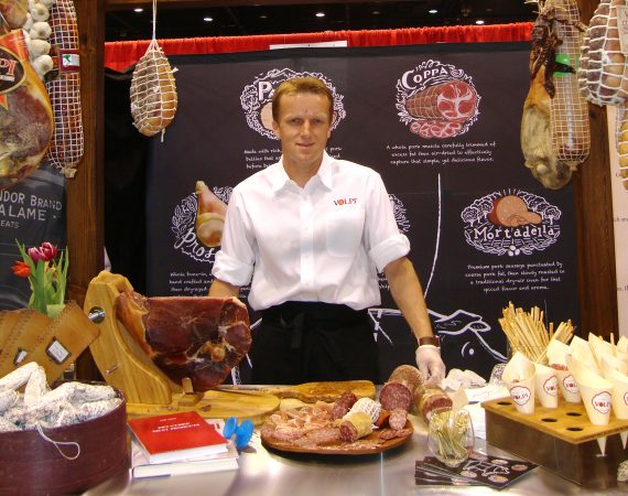 Man standing at a trade show with a display of cured meats